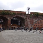 line of cyclists stand astride their bikes leaning to the left, watched by a large audience