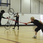 Two dancers leap & lunge, joined by a red silk scarf. In the background a performer walks his tandem on its back wheel