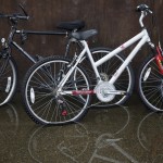 Three bikes leaning against a wall sit in a puddle of water, up to the wheel rims