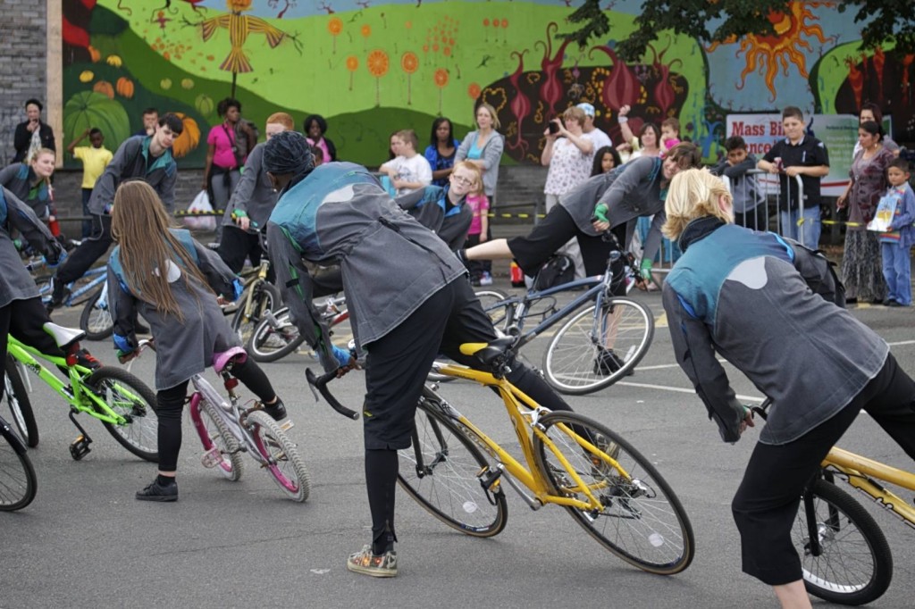a group of performers lean their bikes to the left, with left feet on floor, and right legs raised