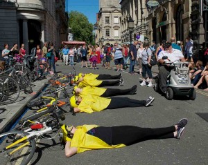 5 performers lie across the road with their heads resting on the saddles of their bikes, which are laid on the ground