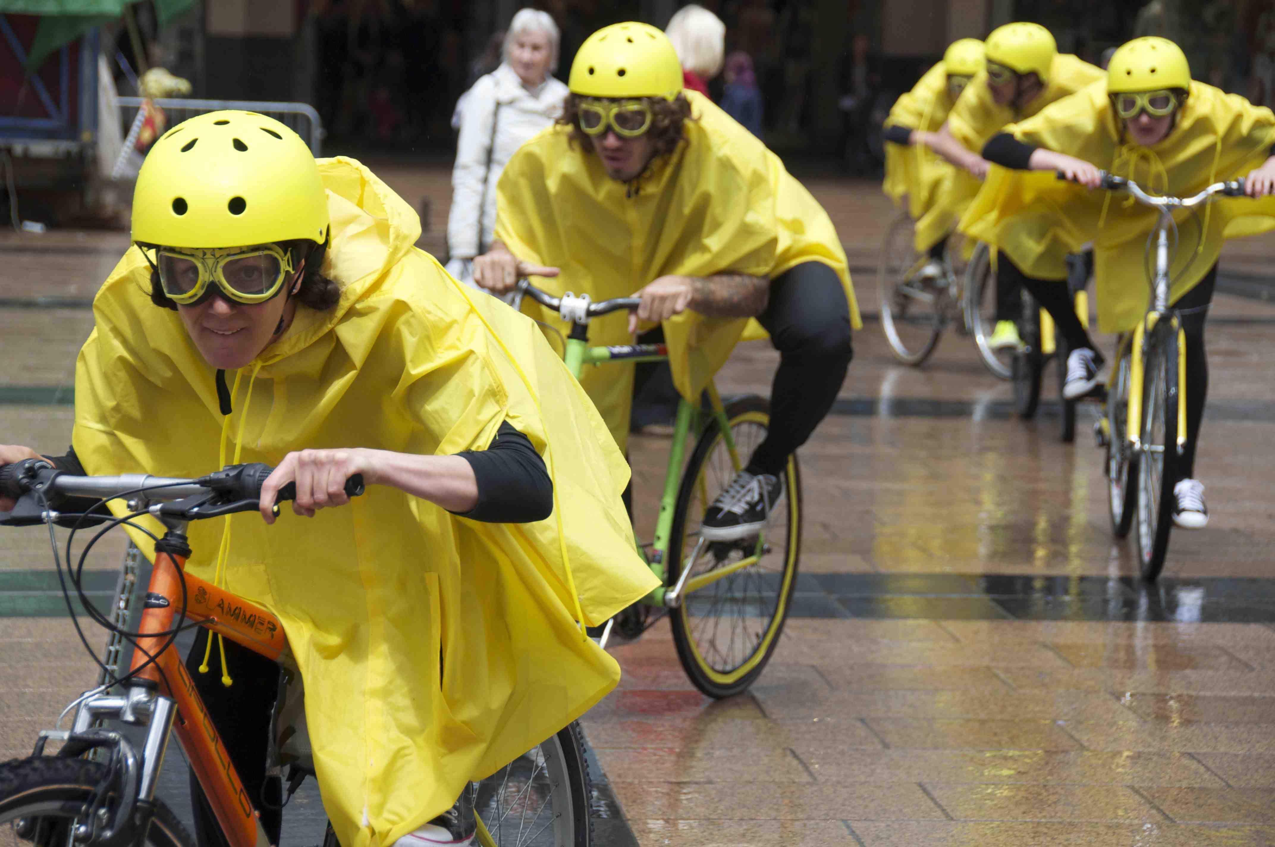five yellow clad cyclings (helment, rain capes & goggles) lean over their handlebars and cycle towards the camera