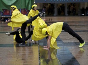 Performers in yellow rain macs, helmets and goggles cluster together forming a human installation