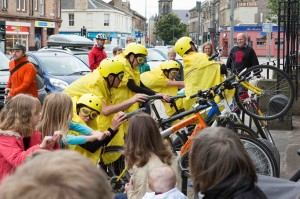 5 yellow cyclists lean over their bikes' back wheels and grab the handlebars, surrounded by people mimicking them without bikes