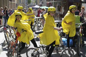 Five performers in yellow helmets, yellow goggles and yellow capes lean against their bikes around a circular table, each looking in different directions and pulling faces