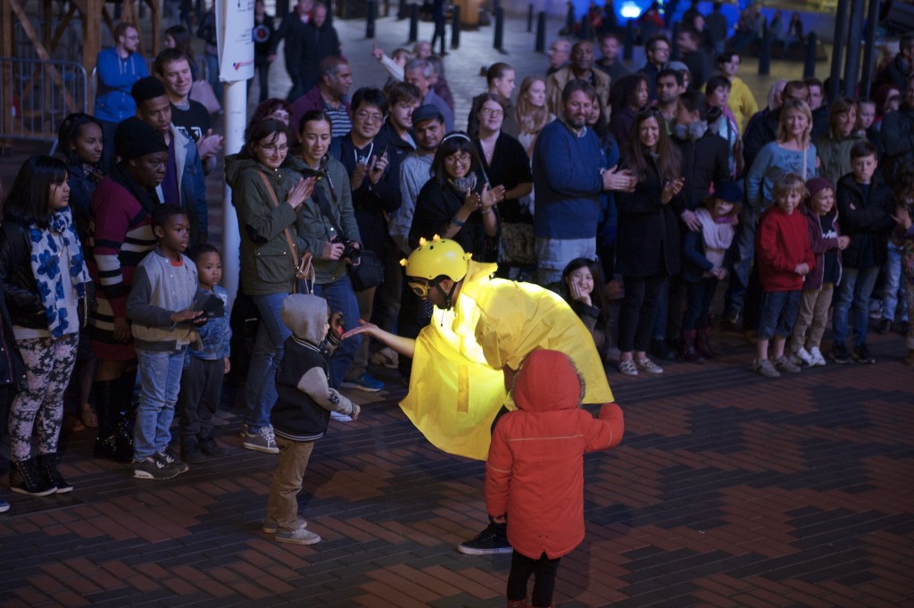 Glowing performer in yellow cape, goggles, with lights on top of helmet, bends down to thank a small boy for participating in the show