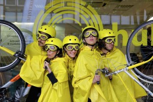 5 performers dressed in yellow rain capes, helmets & goggles huddle together looking in all directions, the outer two holding bicycle handlebars